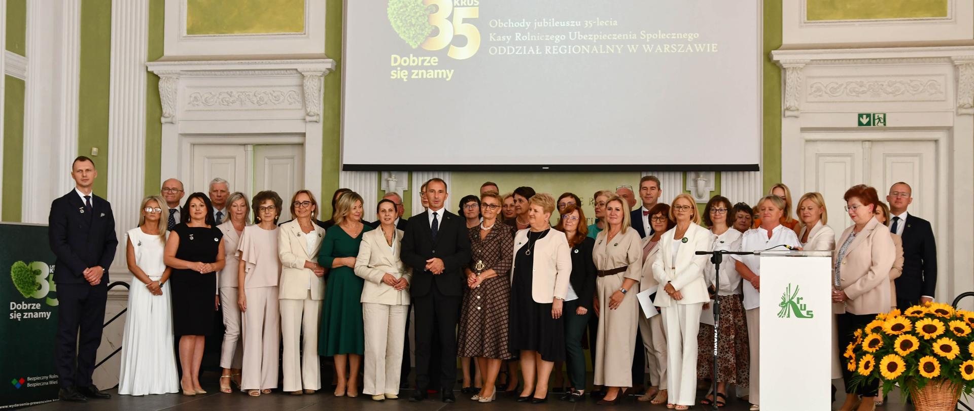 Grupa kilkudziesięciu osób pozuje do wspólnego zdjęcia na scenie auli. Za nimi ekran z napisem 35 lat KRUS . Dobrze się znamy.