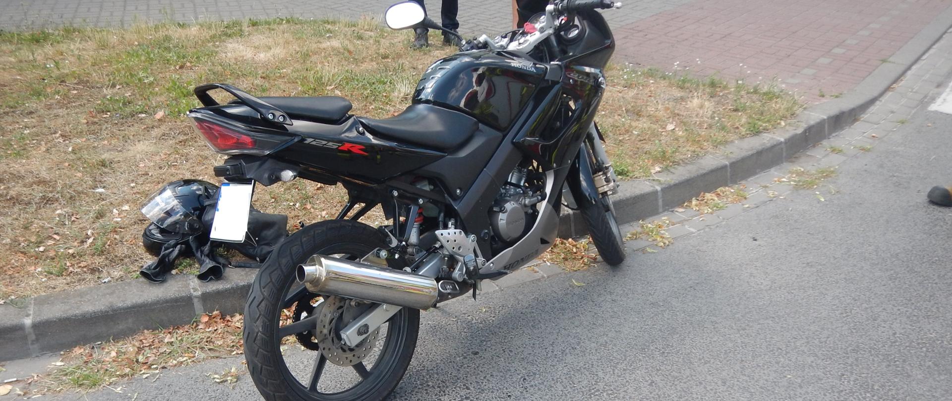 Zdjęcie przedstawia czarny motocykl, który brał udział w zdarzeniu. Obok motocyklu znajduje się kask oraz rękawice kierującego motocyklem. Motocykl znajduje się na skraju drogi.