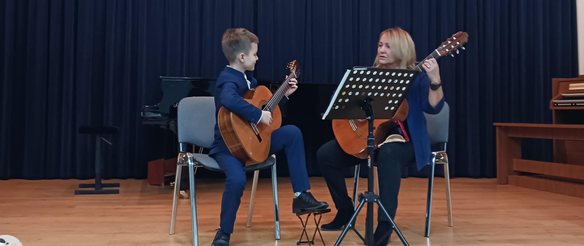 Zdjęcie przedstawia chłopca i kobietę , trzymają gitary , na granatowym tle.