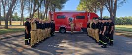 Strażacy stoją na baczność ustawieni na przeciwko siebie w dwóch rzędach. Za nimi stoi wóz strażacki, a dookoła są drzewa. 