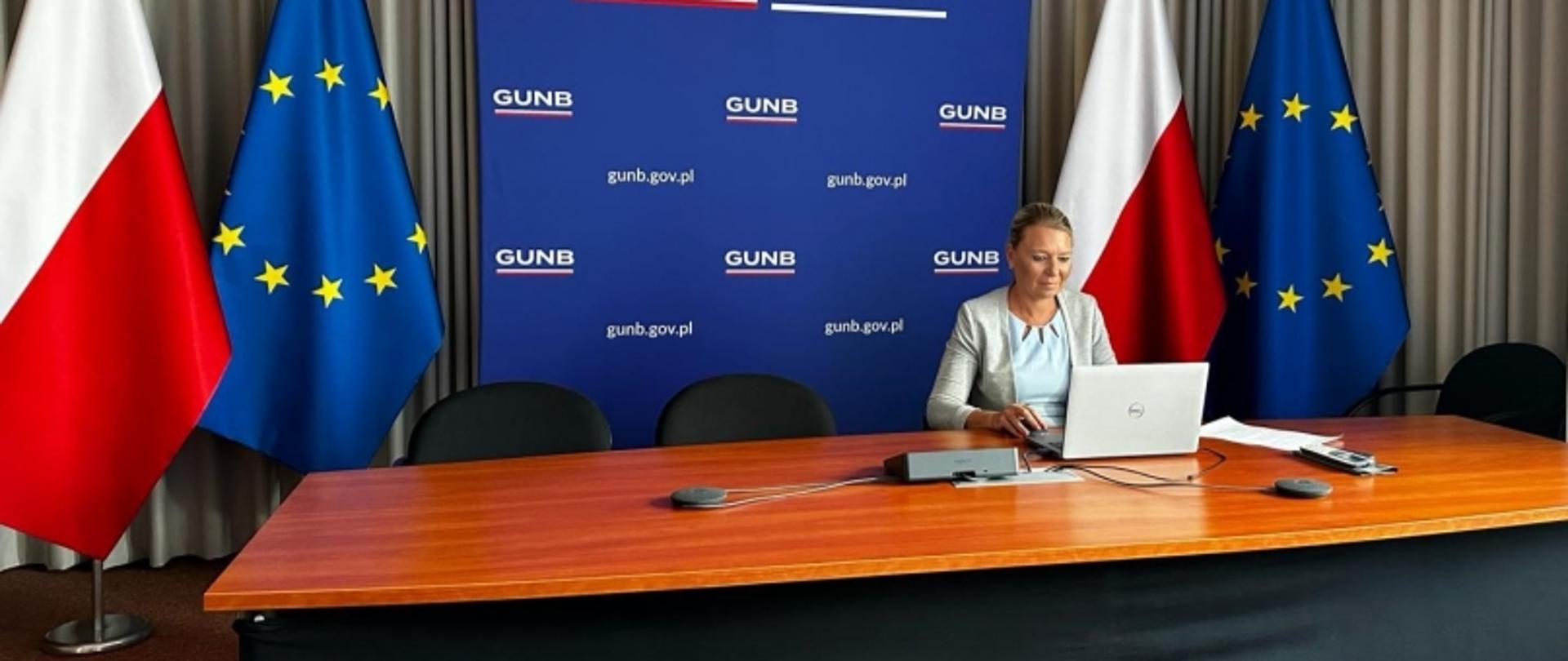 Osoba siedząca przy biurku z laptopem, w tle baner GUNB oraz flagi Polski i UE.
