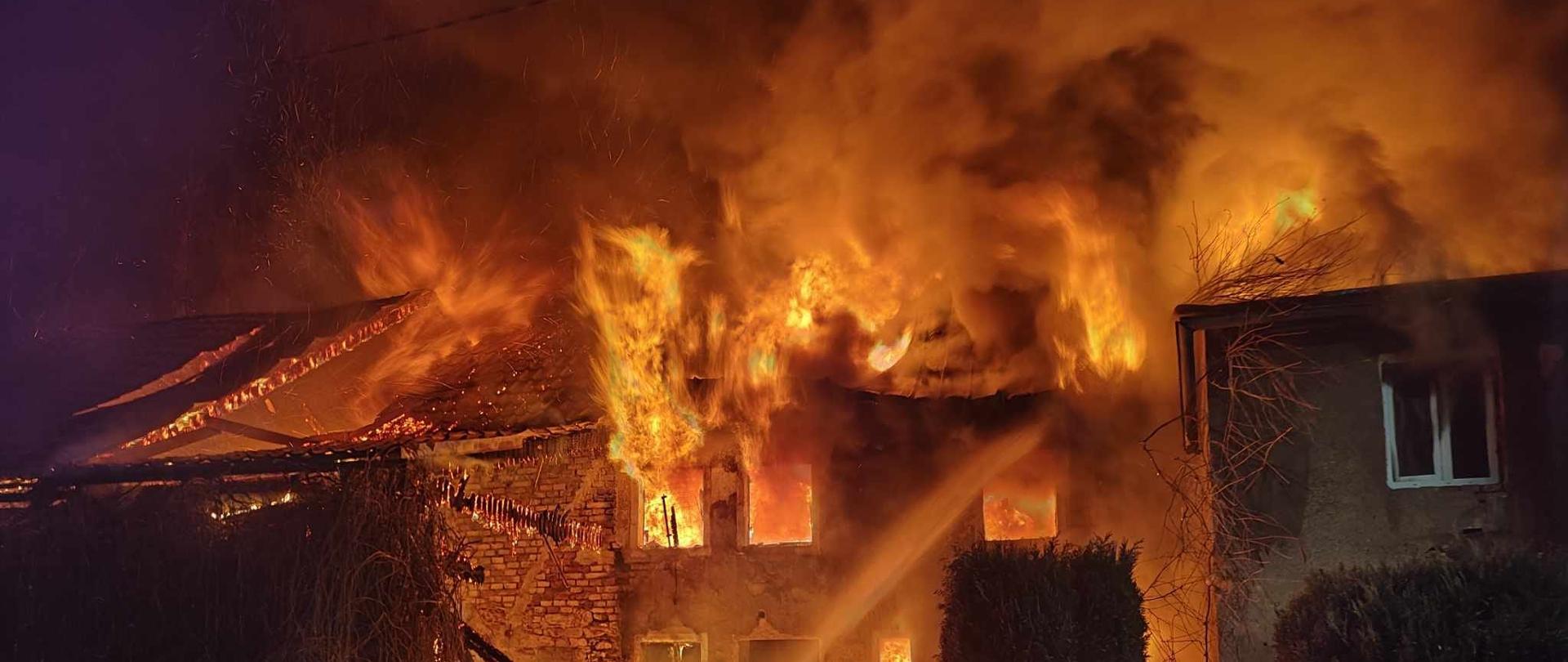 Na zdjęciu widać, palący się budynek mieszkalny i strażaka, który podaje jeden prąd wody na palący się dach.
