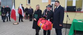 Na zdjęciu widać strażaków oraz ministra stojących przed remizą OSP podczas przekazania samochodu