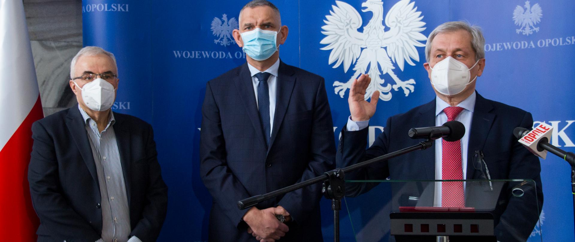 Na zdjęciu trzech mężczyzn. Od prawej: wojewoda opolski, dyrektor USK w Opolu, kierownik Szpitala Tymczasowego w Opolu.
Wojewoda stoi za szklaną mównicą, mówi do zgromadzonych na konferencji dziennikarzy poprzez ustawiony przed nim na stojaku mikrofon. Ma lekko uniesioną prawą dłoń. Drugą opartą ma o mównice. Patrzy w dal. Obok niego, tuż po jego prawej stronie stoi dyrektor USK. To postawy, wysoki mężczyzna. Obok niego stoi kierownik szpitala tymczasowego, na nosie ma okulary optyczne. Mężczyźni ubrani są w granatowe garnitury. Za nimi w tle stoi ścianka także koloru granatowego z napisem WOJEWODA OPOLSKI. Po lewej stronie kadru w dali widać fragment stojącej na stojaku biało-czerwonej flagi. 