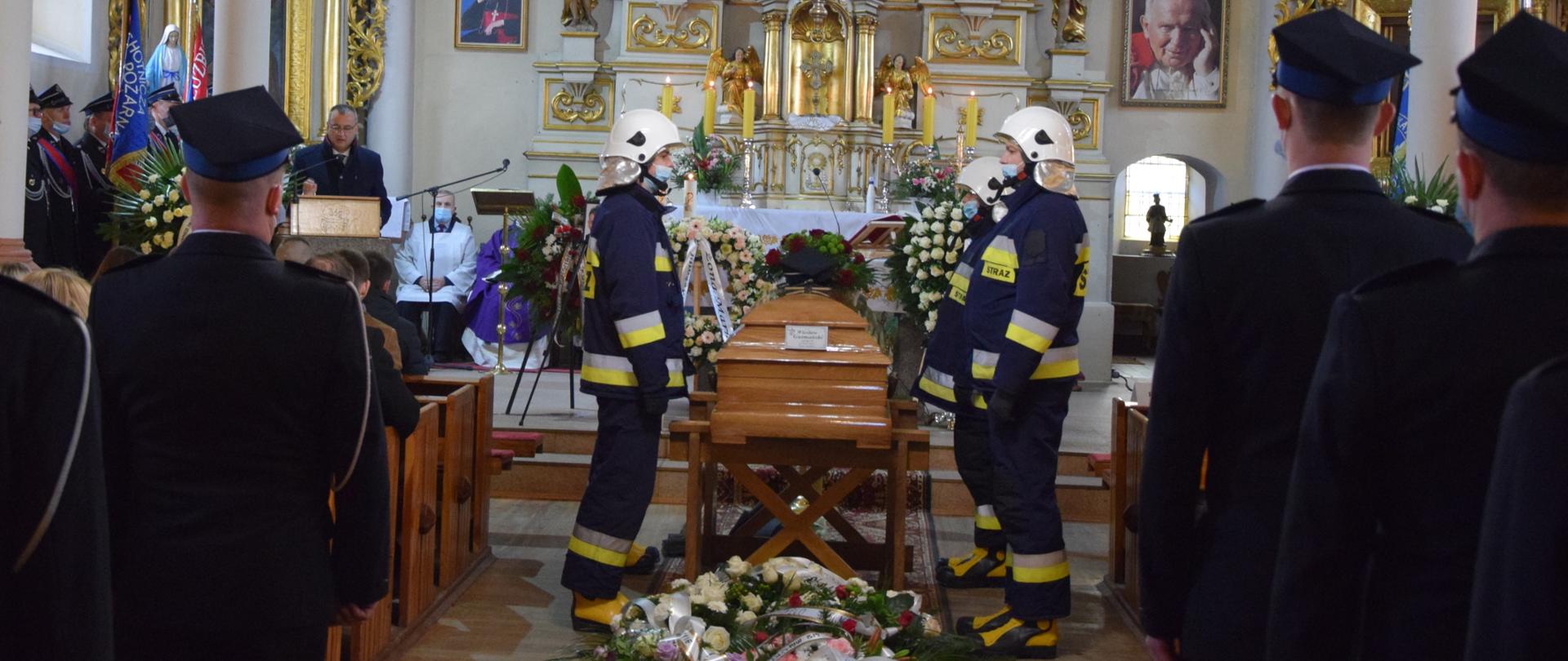 W kościele, przy trumnie asysta honorowa złożona ze strażaków OSP w umundurowaniu bojowym. Przed trumną złożone wieńce pogrzebowe. Po obu stronach strażacy - uczestnicy uroczystej mszy.