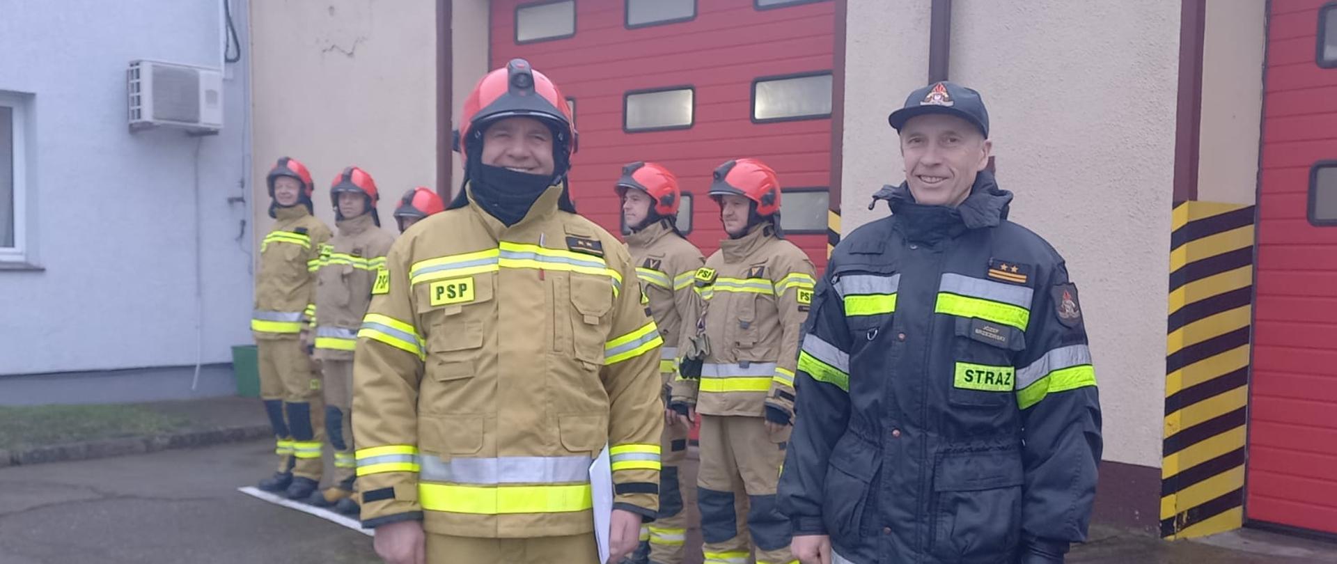 Zdjęcie przedstawia plac przed czerwonymi bramami garażowymi Jednostki Ratowniczo-Gaśniczej w Świdwinie. Przed bramą stoi zmiana służbowa w piaskowych ubraniach specjalnych z czerwonymi hełmami - stoją wzdłuż namalowanej białej linii. Na pierwszym planie znajduje się Komendant Powiatowy PSP w Świdwinie ubrany w czarny mundur. Obok niego stoi dowódca zmiany ubrany w piaskowy mundur i czerwony hełm. W ręku trzyma dokumenty. Panowie się uśmiechają.