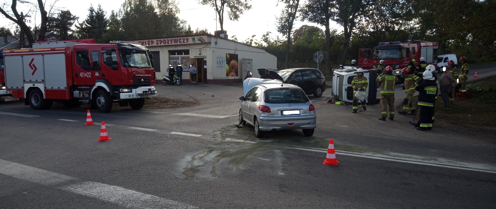 W prawej części zdjęcia strażacy udzielają pomocy medycznej poszkodowanemu mężczyźnie, w środkowej części trwają działania neutralizujące wyciekłe płyny eksploatacyjne