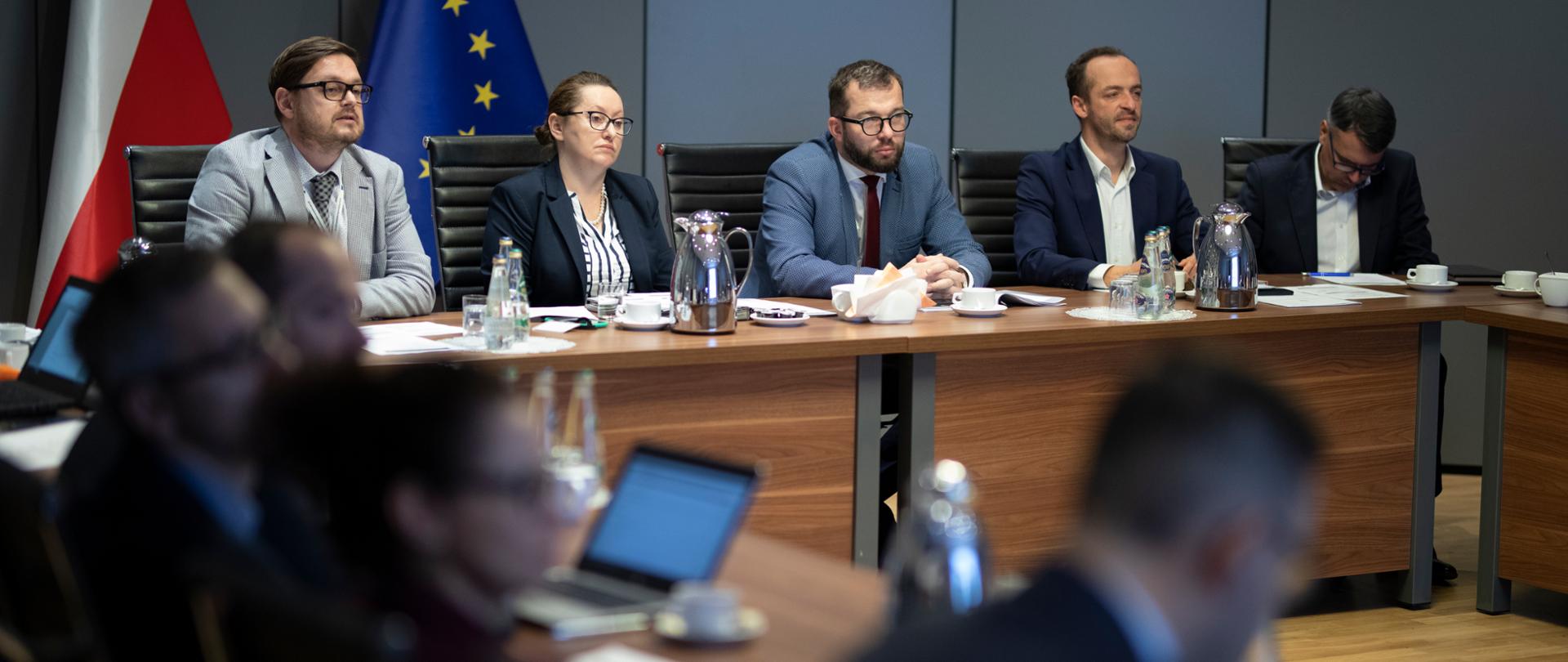 Minister Grzegorz Puda wraz z pozostałymi członkami zespołu siedzi przy stole. Za stołem stoi flaga Polski i Unii Europejskiej. 
