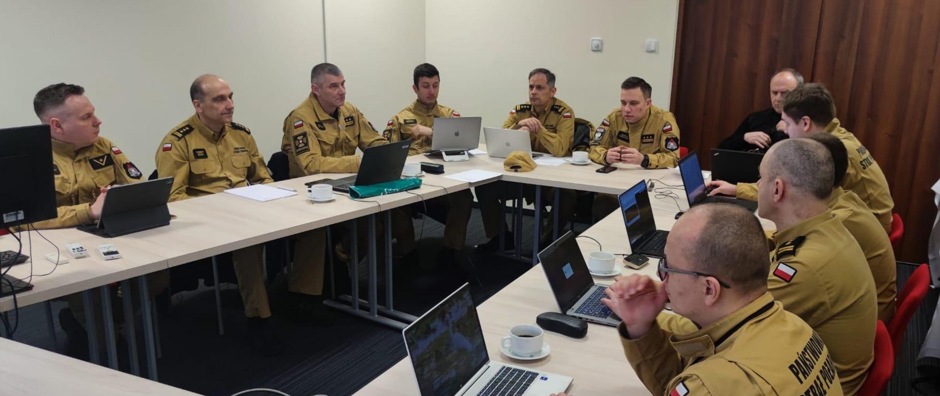 Wewnątrz sali konferencyjnej przy dużym stole w kształcie litery U siedzą strażacy, przed nimi znajdują się laptopy.