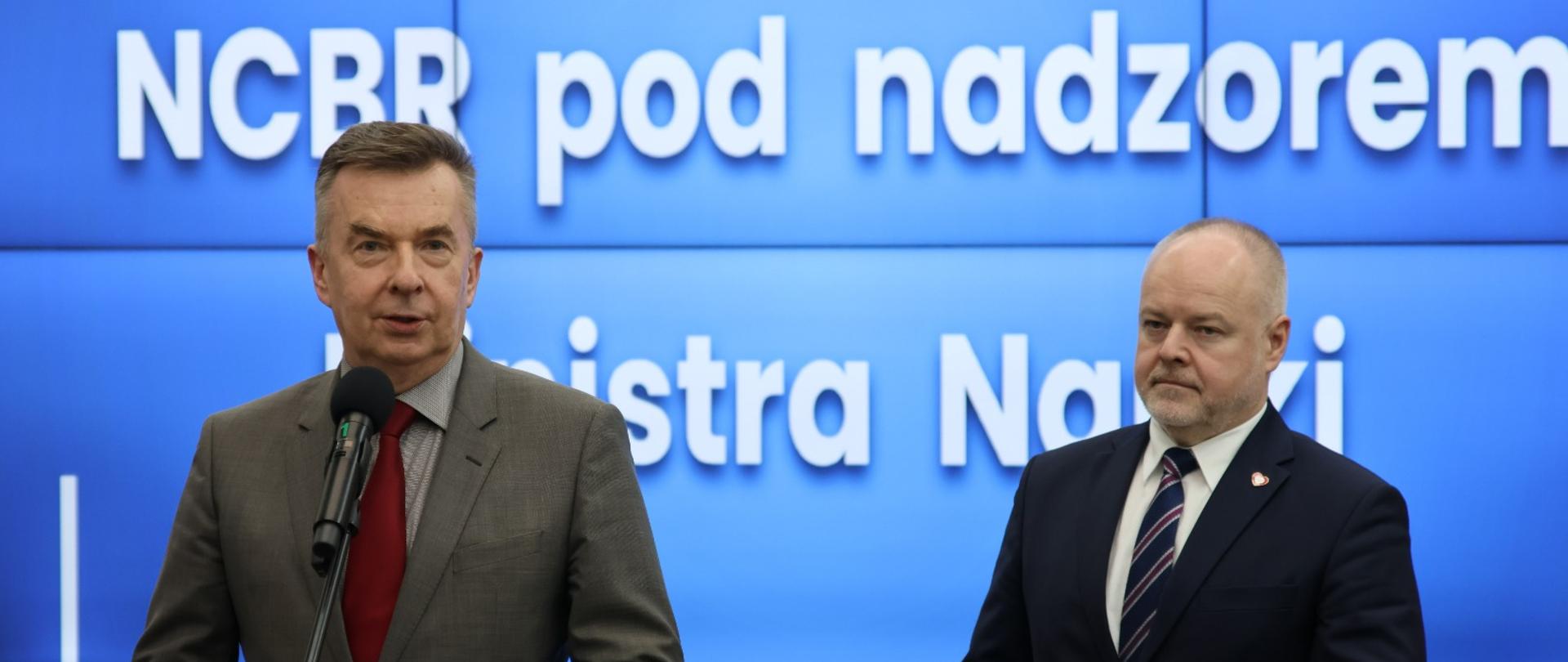 Na tle wbudowanego w ścianę wielkiego ekranu z napisem NCBR pod nadzorem Ministra Nauki stoi minister Wieczorek, który mówi do mikrofonu na stojaku, obok niego wiceminister Gzik.