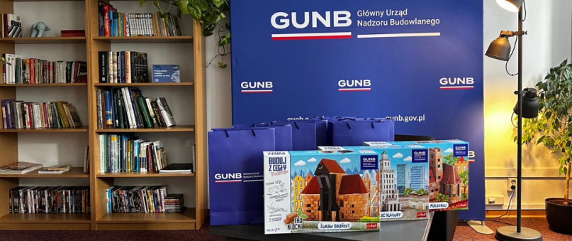 Stół z torbami GUNB i zestawami klocków, na tle regałów z książkami i baneru Głównego Urzędu Nadzoru Budowlanego.