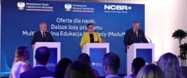 Za trzema mównicami stoją wiceminister Gzik, wiceminister Lubnauer i dyrektor NCBR, za nimi granatowa ścianka z napisem Oferta dla nauki, przed nimi widać siedzących na krzesłach ludzi.