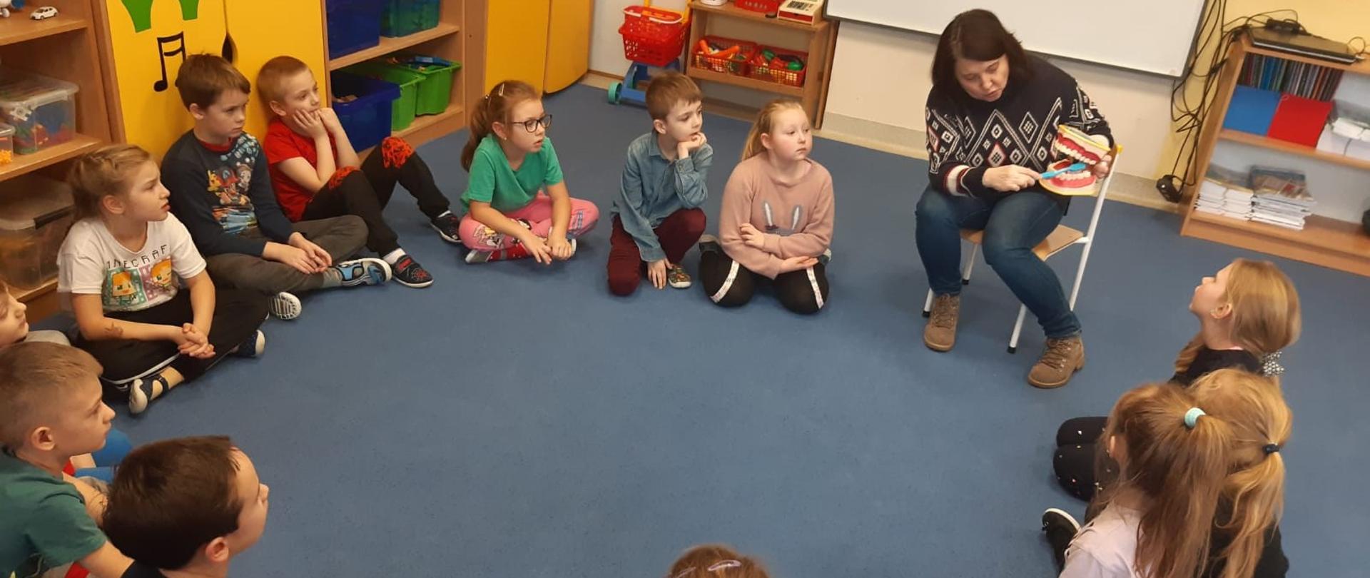 Prelegentka prowadzi instruktaż mycia zębów na modelu szczotki i szczęki otoczona dziećmi siedzącymi w kręgu na dywanie.
