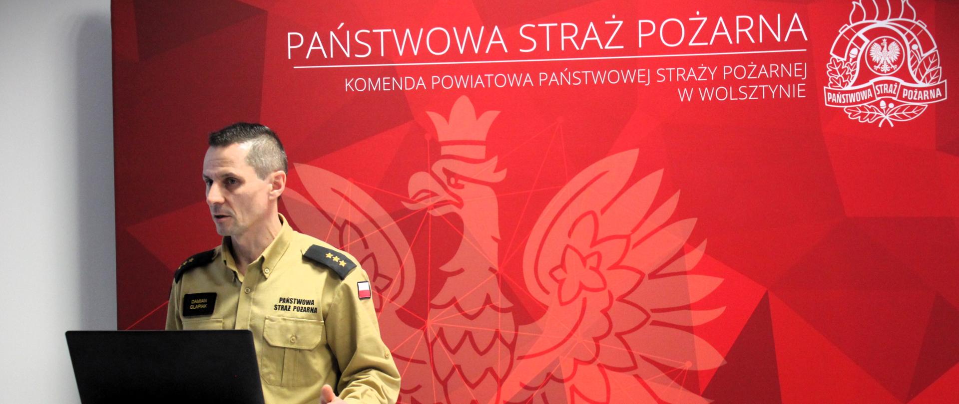 Zdjęcie przedstawia oficera Państwowej Straży Pożarnej w Polsce, który stoi przy podium i prowadzi prezentację lub przemówienie. Ma na sobie mundur galowy w kolorze khaki z naszywką polskiej flagi na ramieniu oraz nazwiskiem na piersi. Na jego pagonach widnieją trzy gwiazdki, co sugeruje stopień oficerski.
Za nim znajduje się czerwone tło z dużym, stylizowanym wizerunkiem orła oraz napisem "PAŃSTWOWA STRAŻ POŻARNA" i "KOMENDA POWIATOWA PAŃSTWOWEJ STRAŻY POŻARNEJ W WOLSZTYNIE", co wskazuje, że zdjęcie zostało wykonane w siedzibie jednostki PSP w Wolsztynie. W prawym górnym rogu widoczne jest również logo PSP.
Przed strażakiem znajduje się laptop podłączony kablem, co sugeruje, że prowadzi prezentację multimedialną lub odprawę dla innych funkcjonariuszy. Jego postawa oraz mimika wskazują na skupienie i zaangażowanie w wystąpienie.

