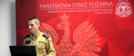 Zdjęcie przedstawia oficera Państwowej Straży Pożarnej w Polsce, który stoi przy podium i prowadzi prezentację lub przemówienie. Ma na sobie mundur galowy w kolorze khaki z naszywką polskiej flagi na ramieniu oraz nazwiskiem na piersi. Na jego pagonach widnieją trzy gwiazdki, co sugeruje stopień oficerski.
Za nim znajduje się czerwone tło z dużym, stylizowanym wizerunkiem orła oraz napisem "PAŃSTWOWA STRAŻ POŻARNA" i "KOMENDA POWIATOWA PAŃSTWOWEJ STRAŻY POŻARNEJ W WOLSZTYNIE", co wskazuje, że zdjęcie zostało wykonane w siedzibie jednostki PSP w Wolsztynie. W prawym górnym rogu widoczne jest również logo PSP.
Przed strażakiem znajduje się laptop podłączony kablem, co sugeruje, że prowadzi prezentację multimedialną lub odprawę dla innych funkcjonariuszy. Jego postawa oraz mimika wskazują na skupienie i zaangażowanie w wystąpienie.
