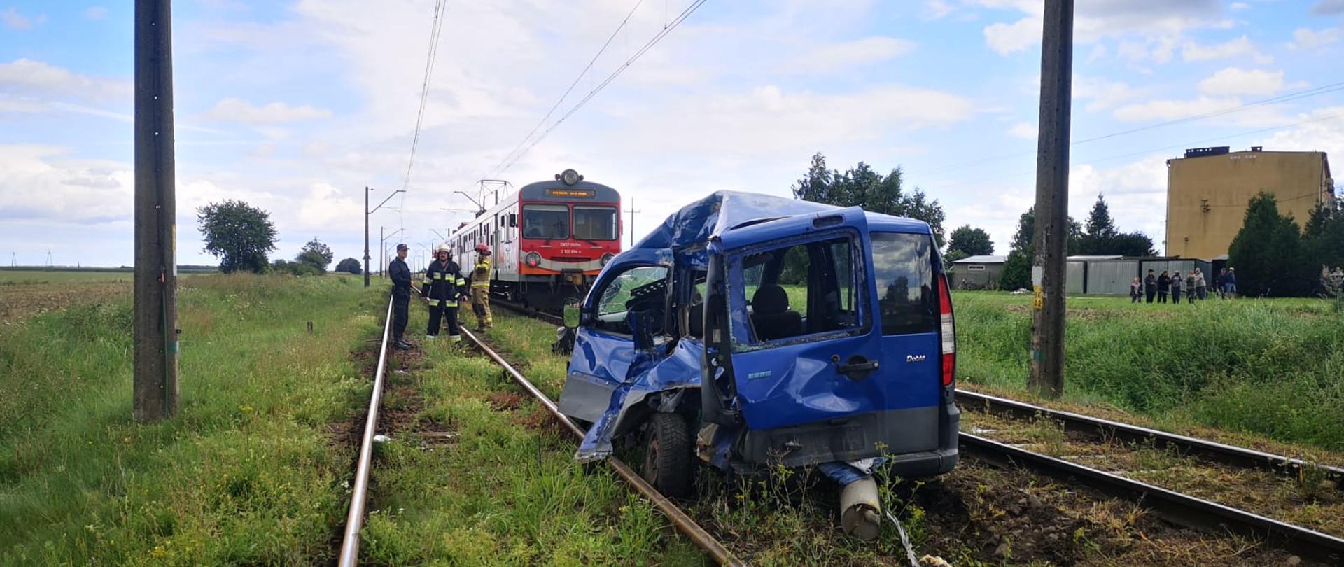 Zdjęcie przedstawie miejsce działań po zderzeniu samochodu osobowego z pociągiem osobowym. Na pierwszym planie widoczny samochód osobowy znacznie zniszczony stojący pomiędzy torami kolejowymi. W oddali trzech strażaków a następnie stojący skład pociągu osobowego.
