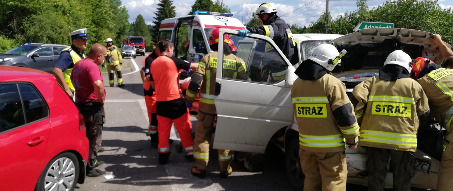 Na zdjęciu widać dwa samochody, jeden osobowy, drugi to bus. Są to pojazdy które brały udział w wypadku drogowym na drodze wojewódzkiej. Po prawej stronie widać znak informujący że na lewo znajduje się droga wiodąca do Arłamowa. Przy busie rozsypany jest sorbent na drodze. Oprócz tego na zdjęciu znajduje się karetka, samochód lekki gaśniczy PSP, strażacy, policjanci, ratownicy medyczni i osoby postronne. 