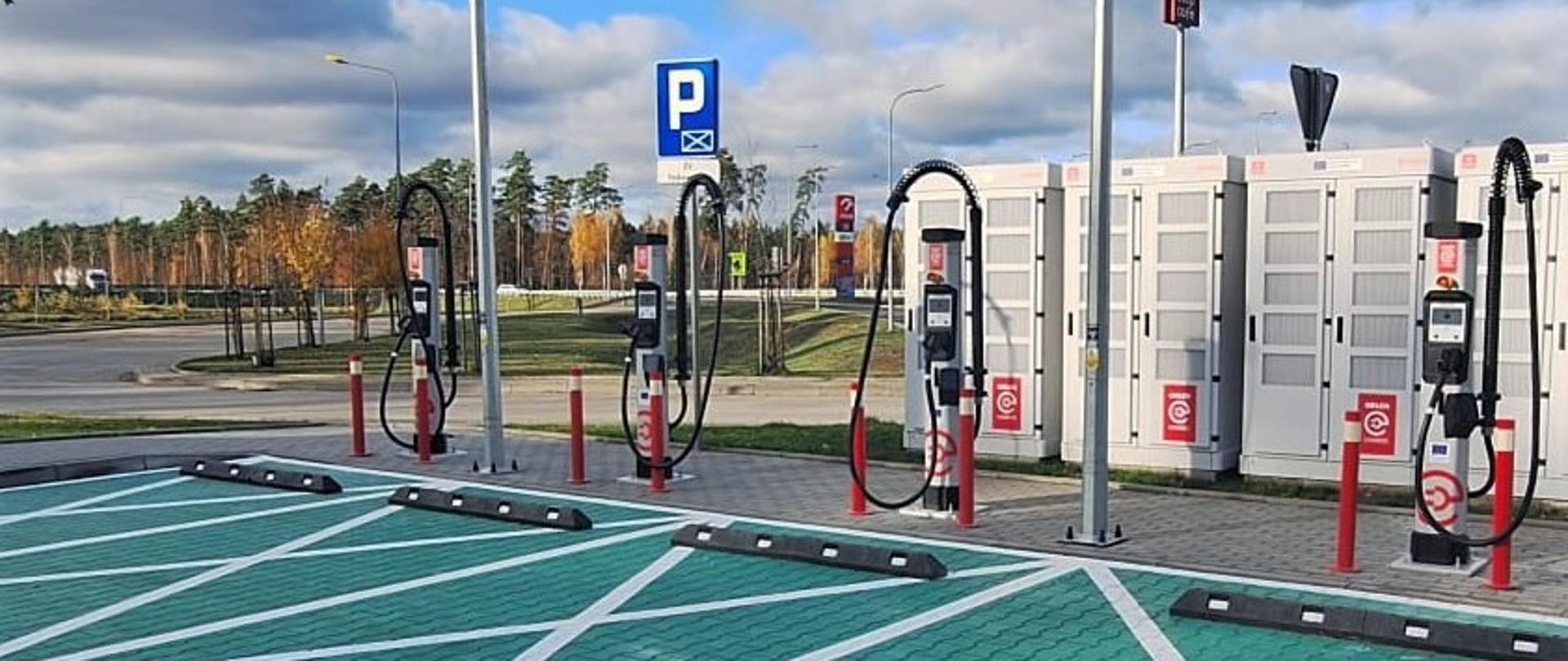 Widok stanowisk do ładowania pojazdów elektrycznych na parkingu. Na pierwszym planie znajdują się zielone miejsca postojowe oznaczone białymi liniami i napisem „EV”. W tle widoczne są cztery słupki ładowarek z przewodami, zabezpieczone czerwono-białymi słupkami. Za nimi znajdują się duże białe moduły techniczne, prawdopodobnie magazyny energii. W otoczeniu widać latarnie, znak parkingu oraz drzewa w jesiennej scenerii pod niebem z chmurami.