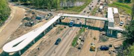 Fotografia lotnicza budowanej kładki dla pieszych nad przyszłą drogą ekspresową S1 przy ulicy Kusocińskiego w Dąbrowie Górniczej. Ustrój nośny jest już prawie gotowy. Na zdjęciu widoczne dwie sekcje rampy przygotowane do zabetonowania. W otoczeniu kładki zgromadzono liczny sprzęt budowlany.