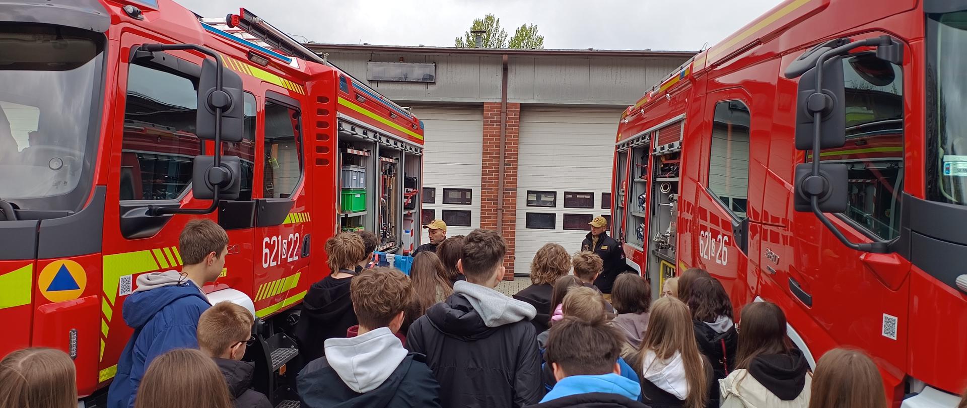 Na zdjęciu widoczna jest grupa młodzieży uczestnicząca w pokazach edukacyjnych zorganizowanych na terenie jednostki straży pożarnej. Uczniowie stoją tyłem do obiektywu, skupiając swoją uwagę na dwóch strażakach w charakterystycznych piaskowych mundurach i czapkach z daszkiem, którzy prezentują wyposażenie techniczne. Wydarzenie odbywa się na placu przed garażami remizy, pod zachmurzonym niebem, co nadaje spotkaniu profesjonalny i roboczy charakter.
Głównym elementem kompozycji są dwa nowoczesne, czerwone wozy strażackie zaparkowane równolegle do siebie, z otwartymi skrytkami sprzętowymi. Na bocznych drzwiach pojazdów widnieją numery operacyjne, a także żółte pasy ostrzegawcze i emblematy obrony cywilnej. Wnętrza ciężarówek wypełnione są specjalistycznymi narzędziami, wężami gaśniczymi oraz pojemnikami technicznymi, które służą strażakom podczas codziennych akcji ratowniczych.
