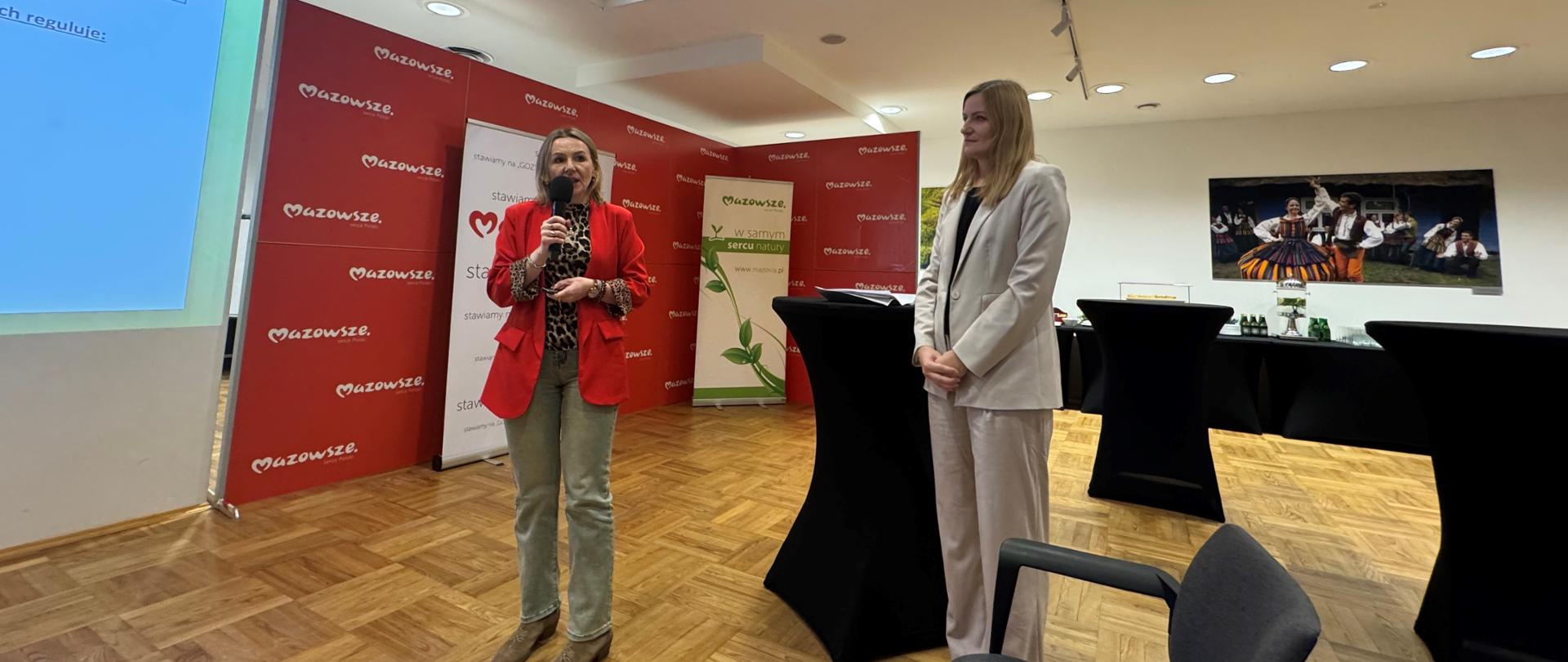 Dwie Inspektorki Wojewódzkiego Inspektoratu Ochrony Środowiska w Warszawie stoją obok siebie podczas prezentacji w sali konferencyjnej. Jedna z nich, ubrana w czerwoną marynarkę, przemawia do mikrofonu, a druga, w jasnym garniturze, stoi obok i słucha. W tle widoczne są banery z logotypem Mazowsze oraz ekran z prezentacją. Sala ma drewnianą podłogę i nowoczesne oświetlenie.
