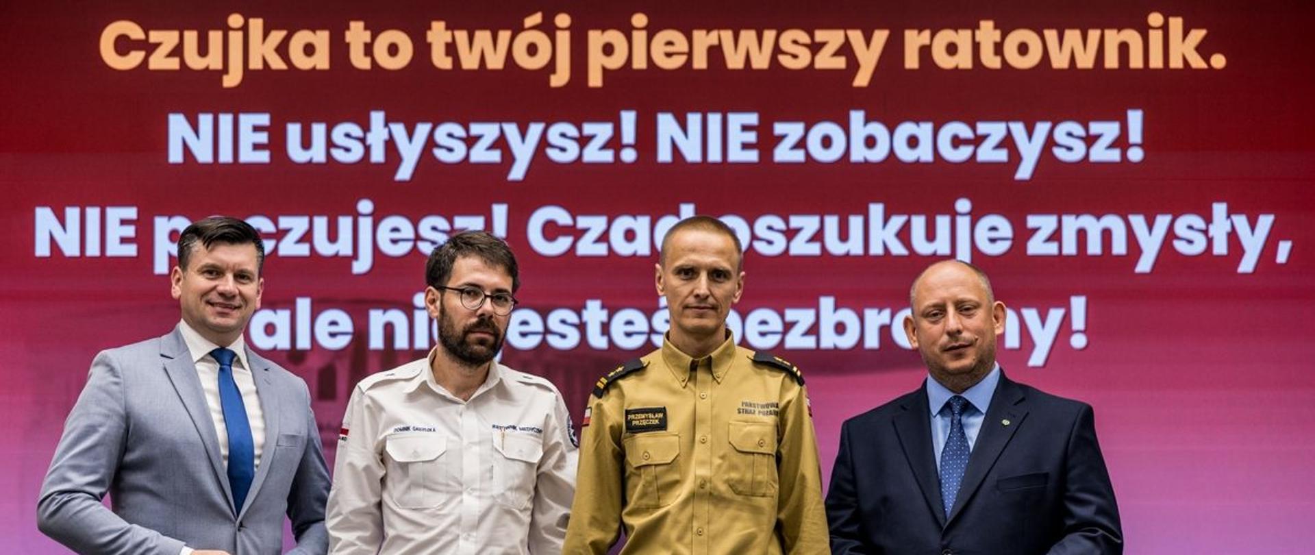 Widok z przodu. Od lewej Marszałek Województwa Małopolskiego, przedstawiciel PRM, Małopolski Komendant Wojewódzki oraz Wojewoda Małopolski na tle napisu "Czujka to twój pierwszy ratownik. NIE usłyszysz! NIE zobaczysz! NIE poczujesz! Czad oszukuje zmysły, ale nie jesteś bezbronny!