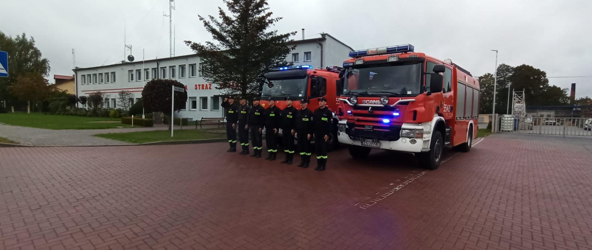Zdjęcie przedstawia dwa wozy ratowniczo gaśnicze stojące przed budynkiem komendy z włączonymi sygnałami świetlnymi i dźwiękowymi oraz strażaków stojących w szeregu na baczność.