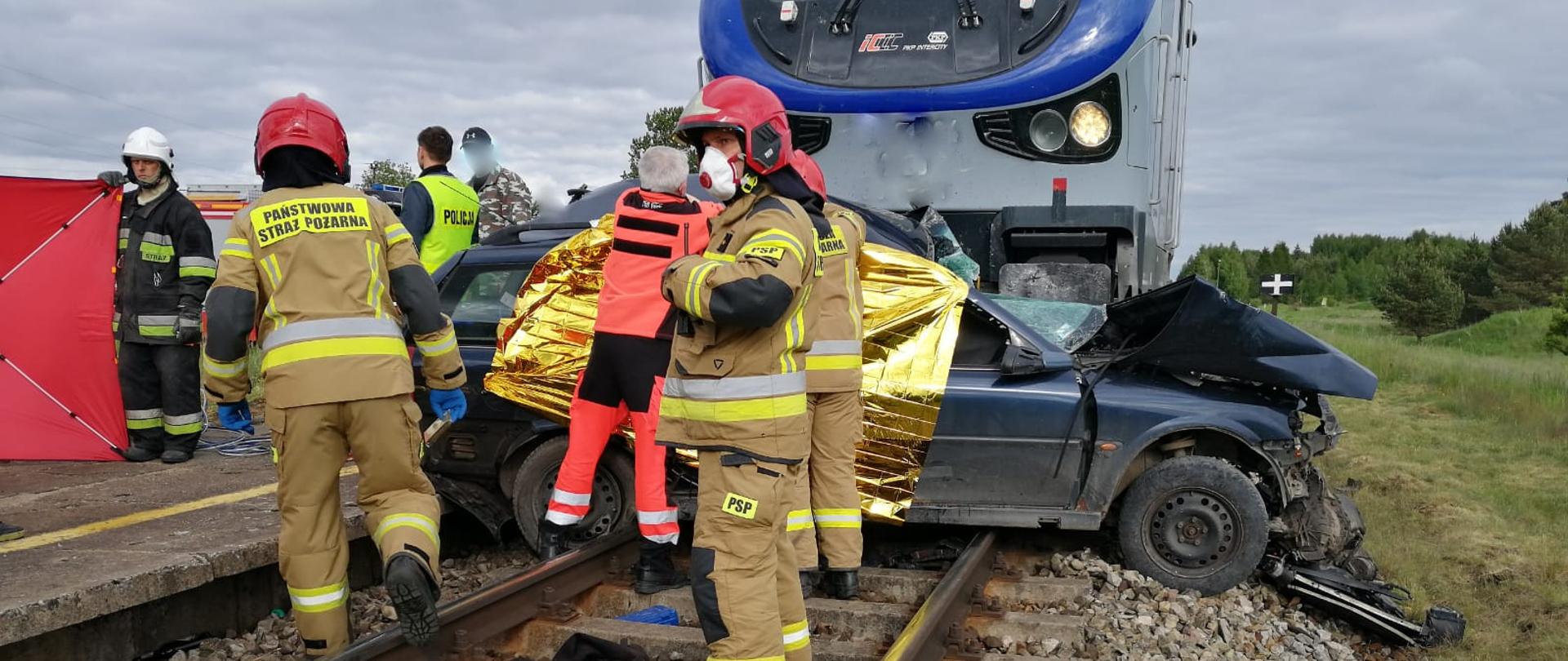 Zdjęcie przedstawia miejsce wypadku. Lokomotywa zmiażdżyła samochód osobowy na torach. No torach stoją funkcjonariusze straży pożarnej w piaskowych ubraniach specjalnych i czerwonych hełmach. Ratownik medyczny w pomarańczowym stroju oraz pozostali mężczyźni. Rozstawiony został czerwony parawan dla poszkodowanego. 