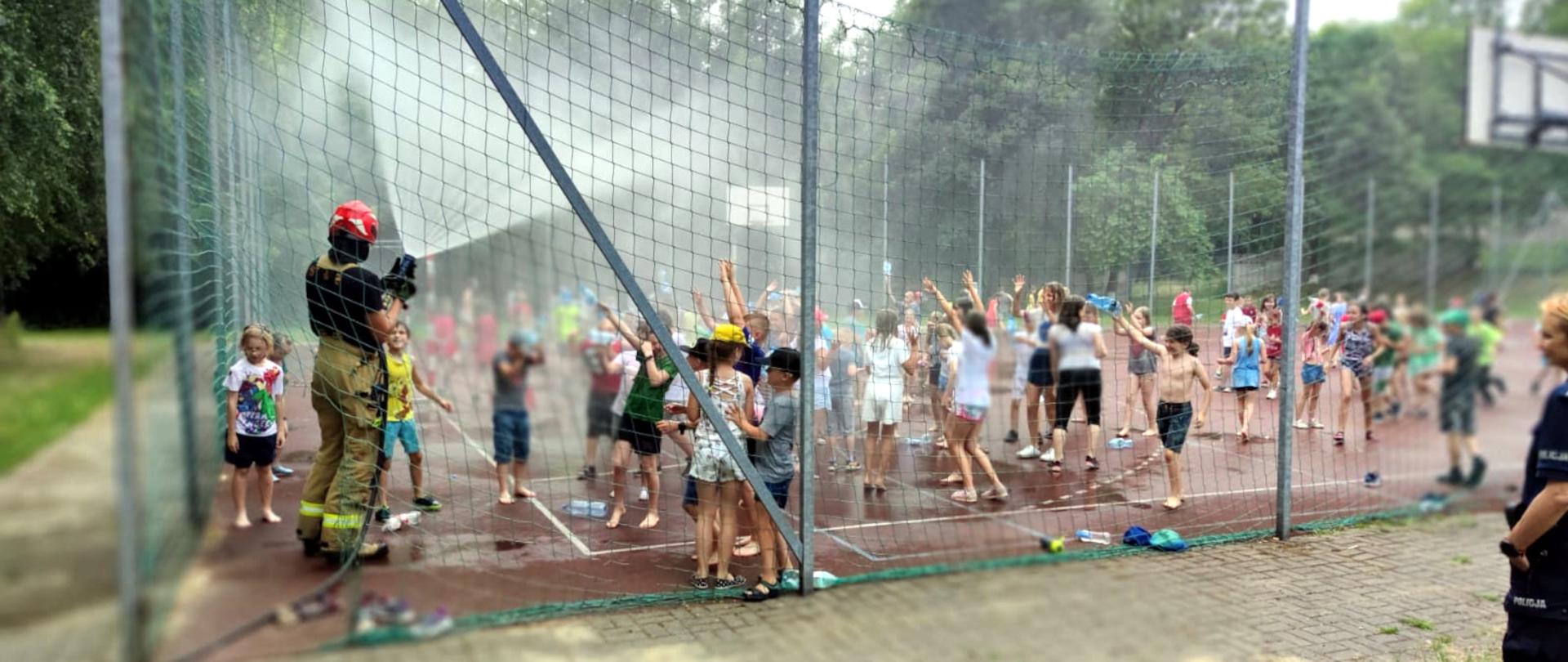 Zdjęcie przedstawia grupę dzieci na boisku szkolnym, które są polewane woda przez strażaka.