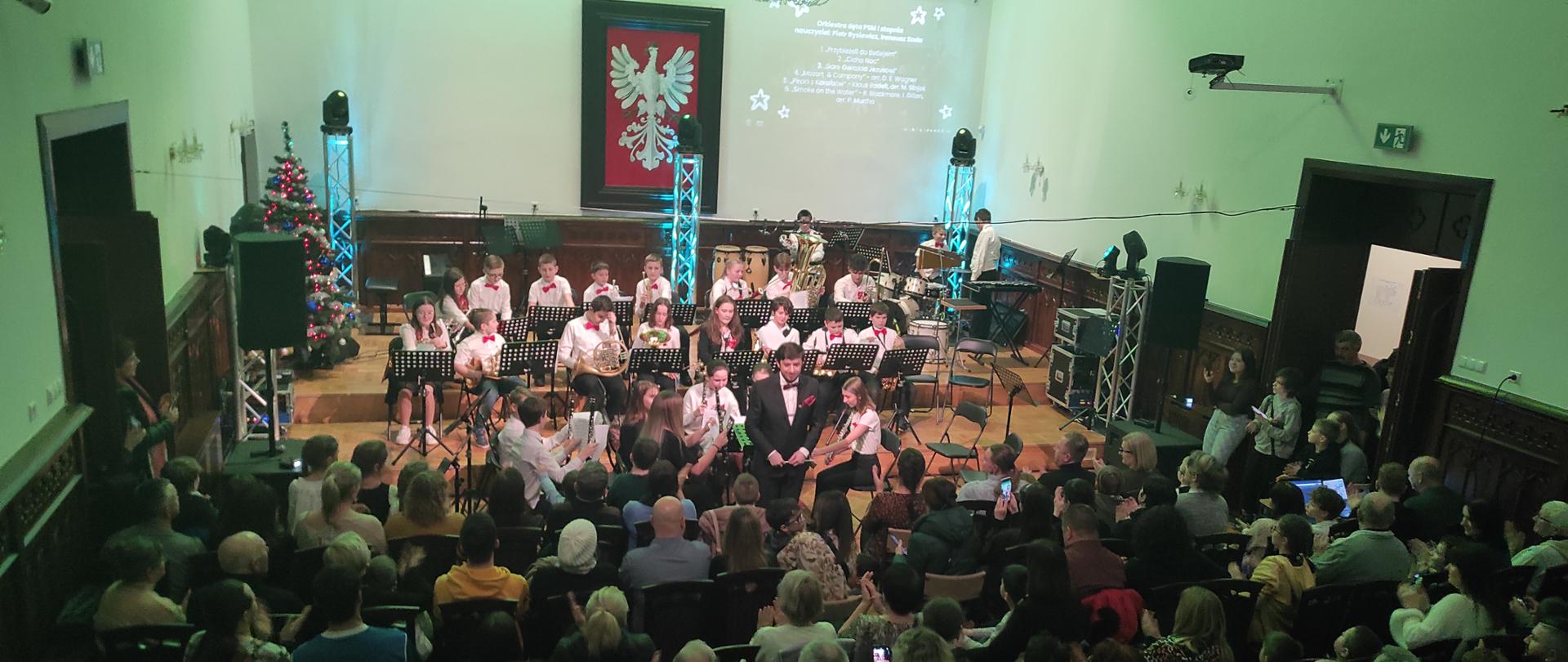 zdjęcie pokazuje Salę Królewską w całej okazałości w zielonkawych poświatach, wypełnioną po brzegi publiczność oraz, na scenie, orkiestrę dętą PSM I stopnia w MIelcu, z dyrygentem na czele