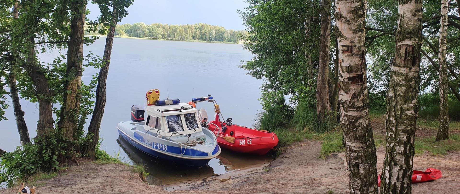 Na zdjęciu widzimy zacumowane do brzegu dwie łodzie ratownicze. Łódka po prawej to łódź strażacka koloru czerwonego, natomiast po lewej stronie kadru to łódź policyjna koloru biało niebieskiego. W tle widzimy jezioro oraz drzewa. 