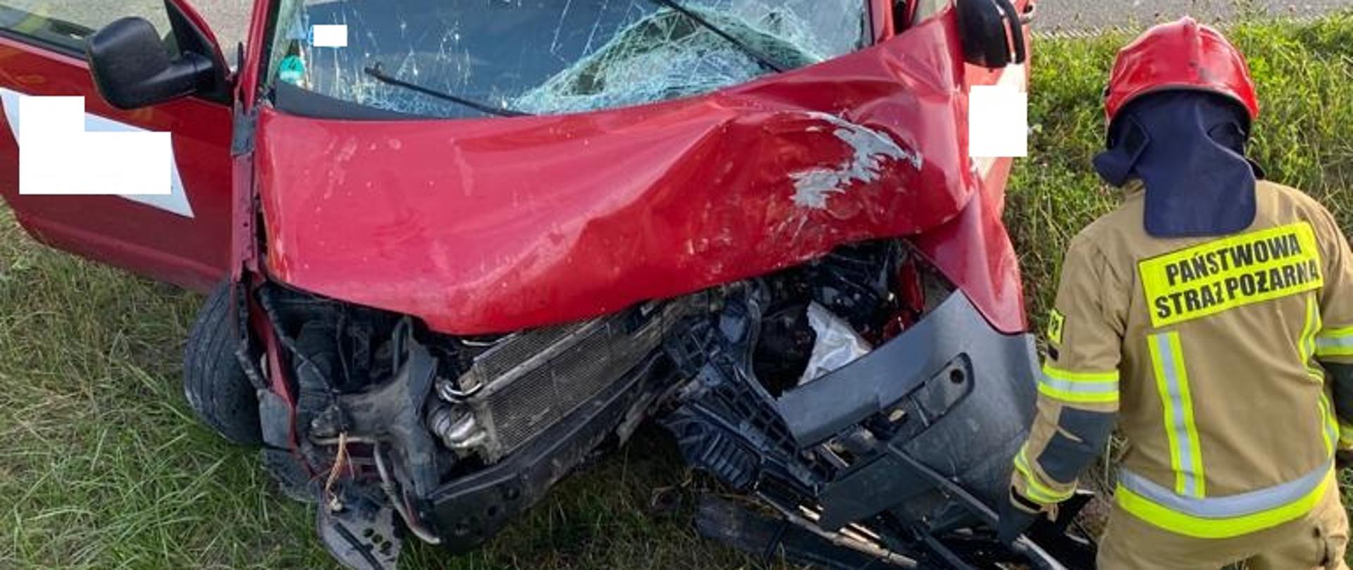 Czerwony samochód bus stoi w rowie po uderzeniu w drzewo, ma otwarte drzwi, przed samochodem stoi strażak. 