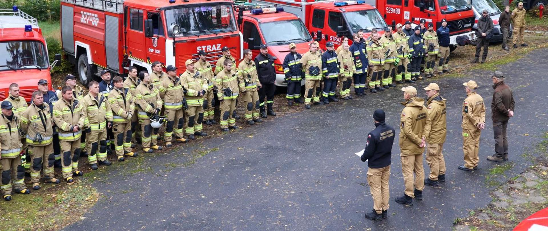 Strażacy ochotnicy stoją przed swoimi samochodami oraz przed kadrą dowódczą rozpoczynającą warsztaty ratownicze.