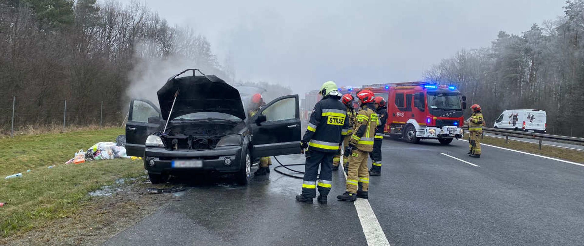 Na zdjęciu znajdują się strażacy, wóz strażacki oraz wrak spalonego pojazdu. Sytuacja ma miejsce na pasie autostrady