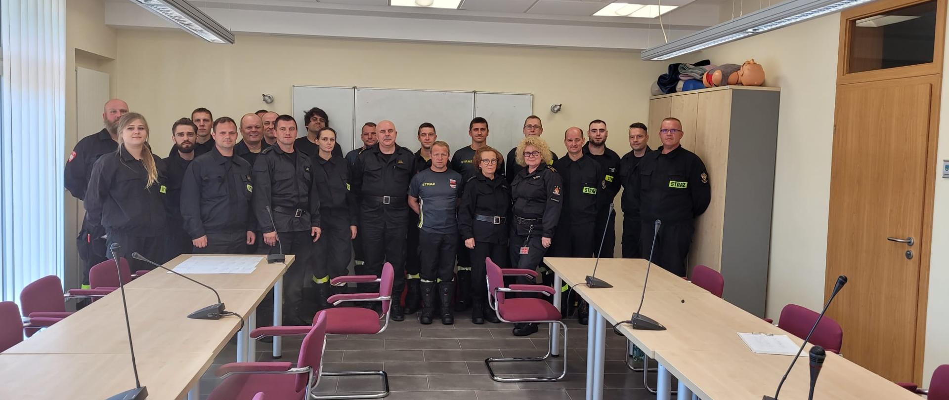Na wspólnym zdjęciu grupa druhów jednostek ochotniczych straży pożarnych przystępujących do egzaminu kwalifikowanej pierwszej pomocy razem z przedstawicielami komendy miejskiej państwowej straży pożarnej w Tychach oraz komendy wojewódzkiej państwowej straży pożarnej w Katowicach. Zdjęcie w pomieszczeniu sali sztabowej na tle białej tablicy ściennej.