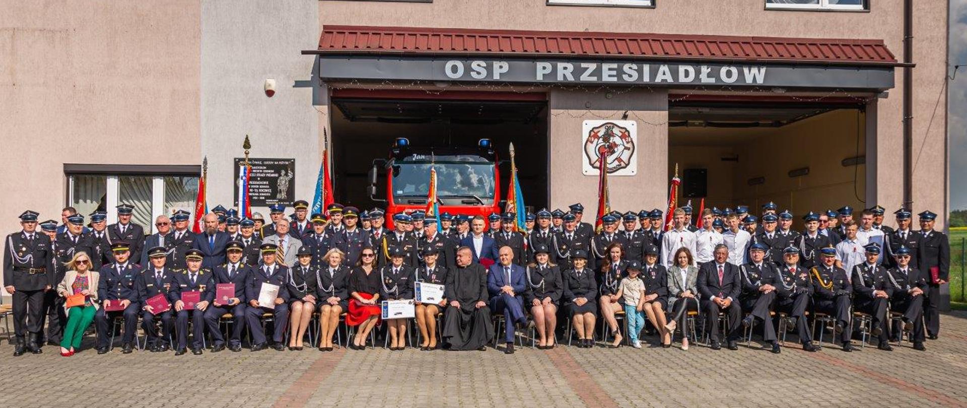 wszyscy uczestnicy uroczystości pozują do zdjęcia na tle budynku remizy OSP Przesiadłów