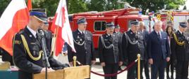 Zdjęcie przedstawia przemawiającego podczas uroczystego apelu Komendanta Powiatowego. Komendant stoi opierając się o mównicę z godłem na froncie. Za nim powiewają flagi. Po prawo stoją zaproszeni goście – większość w mundurach galowych. Za nimi pojazdy pożarnicze – zabytkowe i współczesne.
