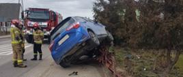 Na zdjęciu widać auto osobowe, które wjechało w ogrodzenie i na nim zawisło, obok stoją strażacy. W tle samochód ratowniczo - gaśniczy.