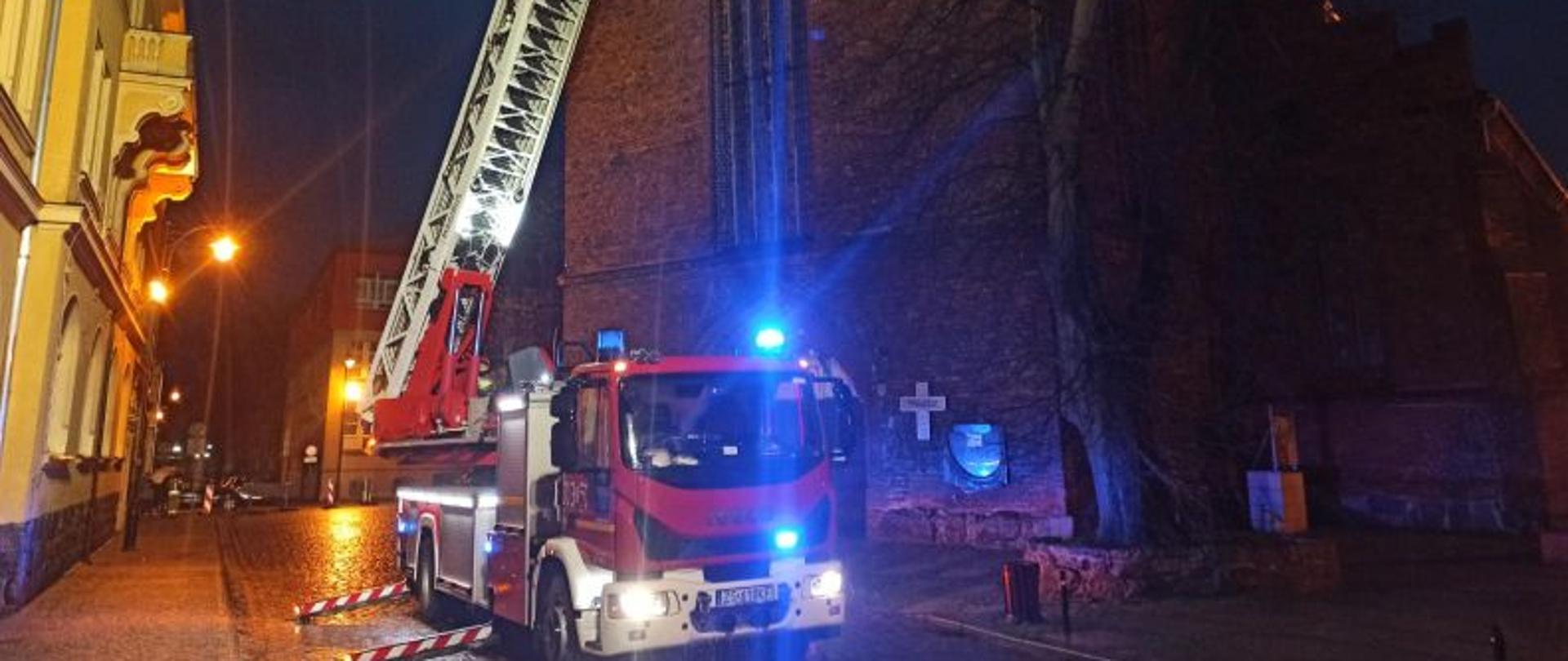 Zdjęcie wykonane w godzinach nocnych. Oświetlone sztucznym światłem z podnośnika straży pożarnej o pobliskich lamp. W centralnym punkcie zdjęcia podnośnik hydrauliczny w trakcie rozkładania. W tle kościół. Auto ma rozłożone podpory i trwa rozkładanie drabiny z koszem