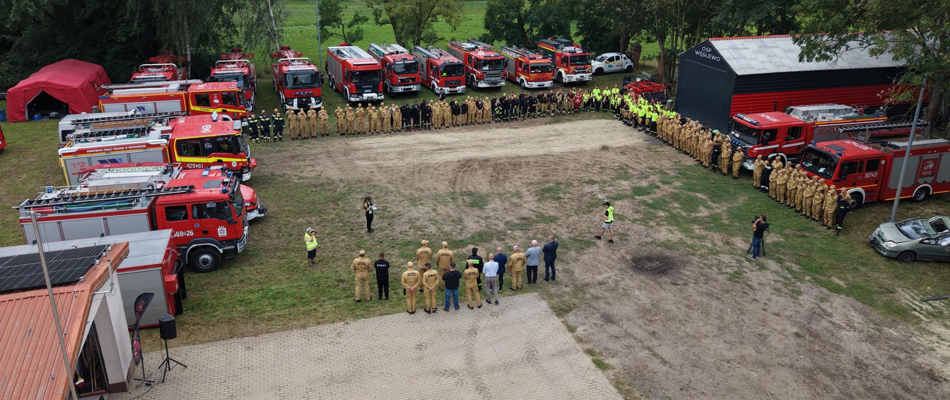 Zdjęcie przedstawia duże zgromadzenie strażaków i pojazdów pożarniczych na terenie jednostki Ochotniczej Straży Pożarnej. Wokół placu ustawionych jest kilkanaście czerwonych samochodów ratowniczo-gaśniczych różnych typów. W centrum oraz wzdłuż pojazdów stoi duża grupa strażaków w umundurowaniu, tworząc półokrąg. W tle widoczny jest budynek oznaczony napisem „OSP WĘGLEWO”, a za nim rozległe tereny zielone i pola uprawne. Zdjęcie wykonano z góry, prawdopodobnie z użyciem drona.