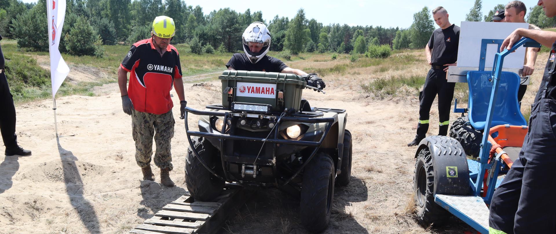 Na zdjęciu widać pojazd typu quad w trakcie instruktażu przejazdu przez przeszkodę powodującą silne pochylenie pojazdu. Na pierwszym planie widać instruktora oraz uczestnika szkolenia prowadzącego quada. Po bokach widać uczestników szkolenia.