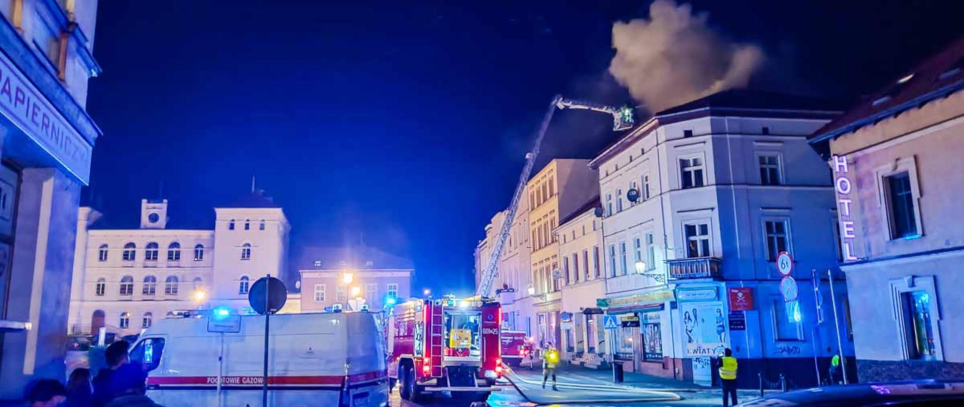 Na zdjęciu widać z poddasza wydobywa się dym na jezdni samochody ratowniczo - gaśnicze i podnośnik z którego podawany jest prąd wody 