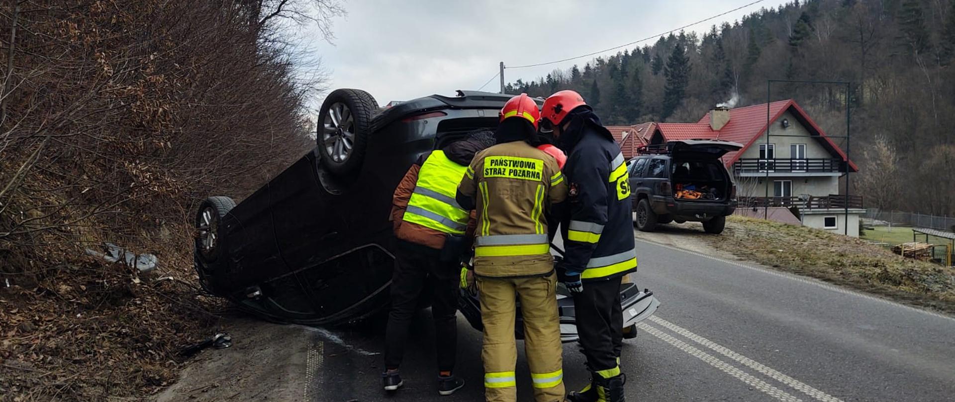 Strażacy obok samochodu osobowego znajdującego się na dachu.