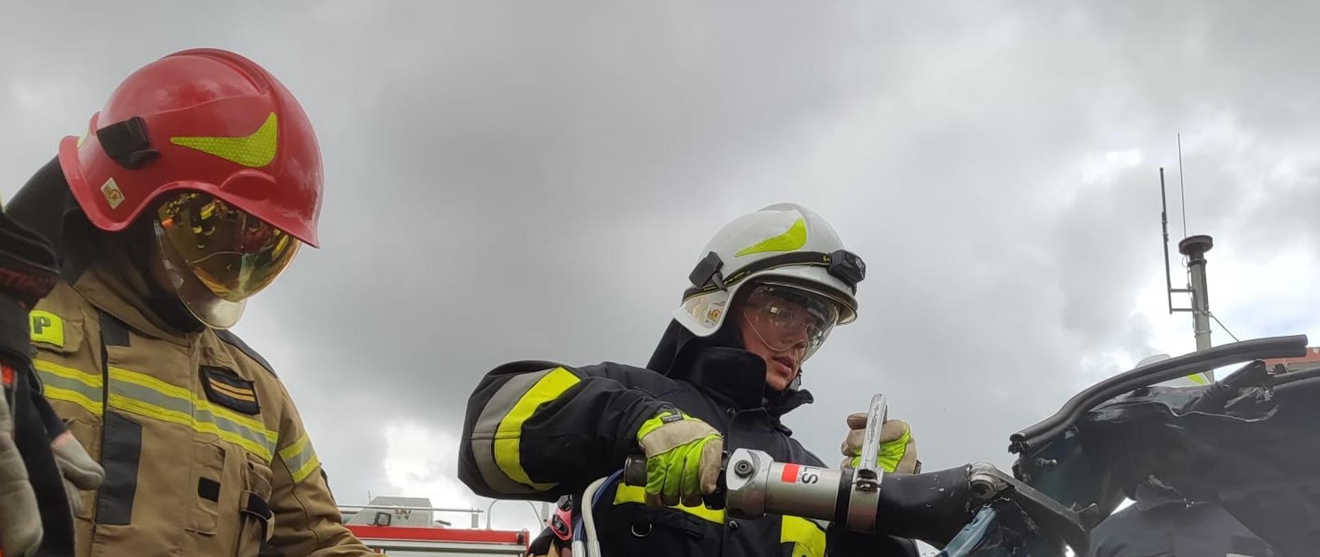 Strażak ćwiczy techniki cięcia pojazdu przy pomocy narzędzi hydraulicznych. Dodatkowo obserwuje go 2 strażaków. 