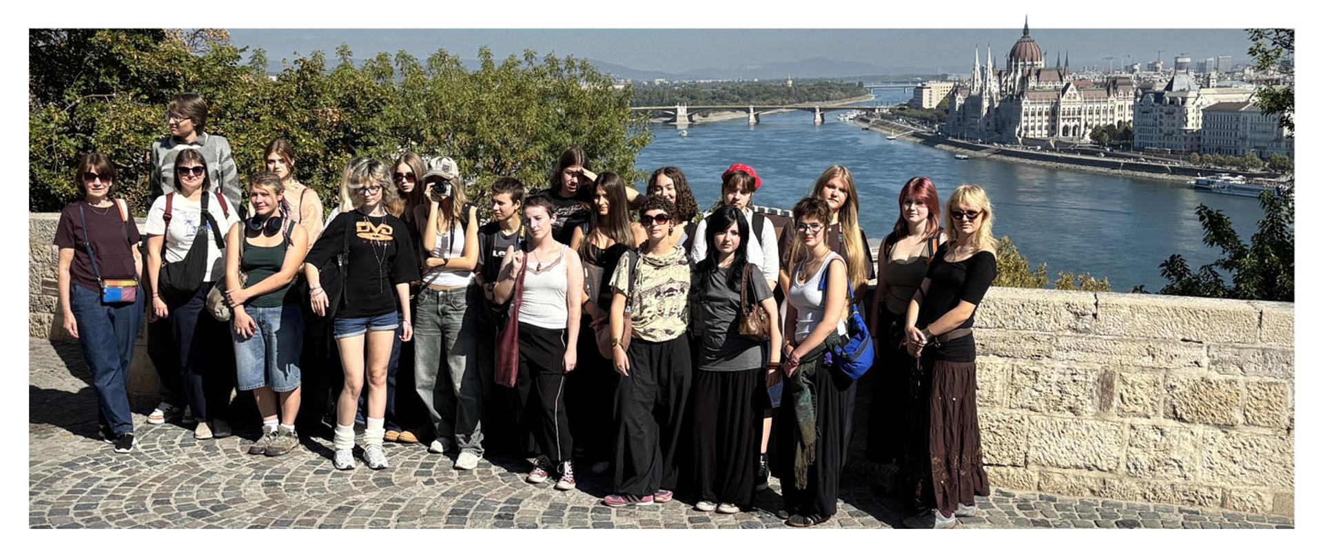 Zdjęcie grupowe uczniów i opiekunów na punkcie widokowym w Budapeszcie. W tle rozpościera się panorama Dunaju oraz charakterystyczny budynek węgierskiego Parlamentu.