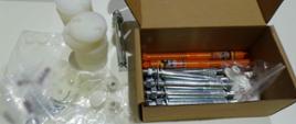 Na białej powierzchni leżą plastikowe elementy, śruby oraz narzędzia częściowo włożone do otwartego kartonu; woreczki z drobnymi częściami.
