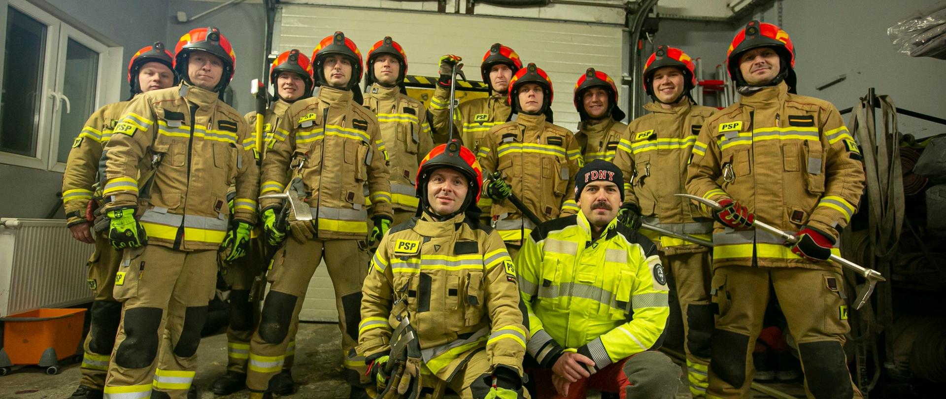 Grupa strażaków w mundurach bojowych i czerwonych hełmach. Pozują do zdjęcia w garażu bojowym.
