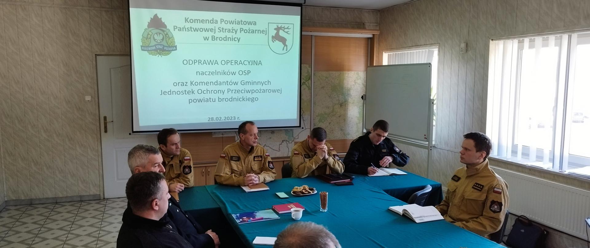 Zdjęcie przedstawia funkcjonariuszy PSP w Brodnicy oraz druhów OSP siedzących przy stole podczas narady. W tle prezentacja wyświetlana na ekranie.