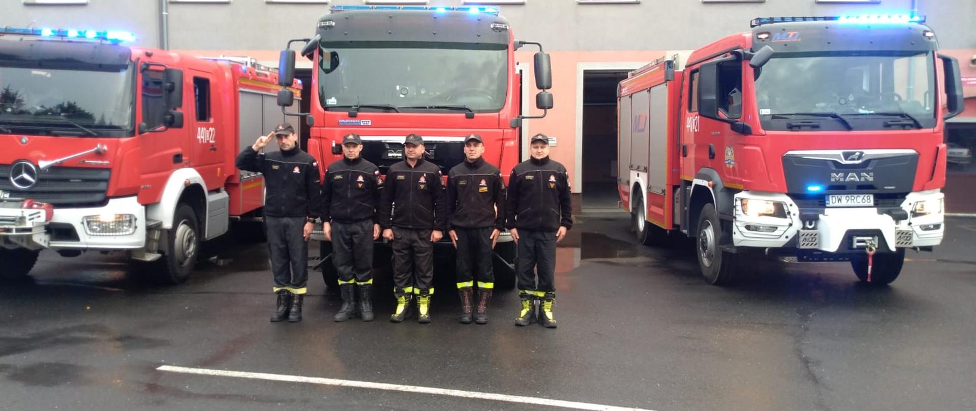 Obraz przedstawia strażaków JRG Jawor podczas minuty ciszy dla zmarłego strażaka. Strażacy w umundurowaniu koszarowym stoją na tle pojazdów pożarniczych i budynku komendy. Pora dzienna.