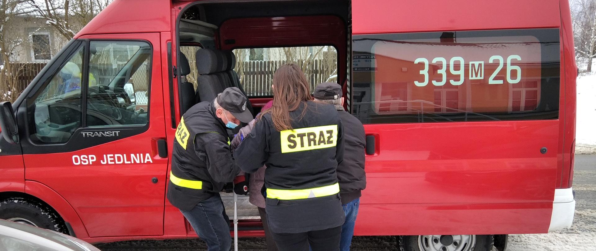 Na zdjęciu widoczni są strażacy z OSP Jedlnia, którzy pomagają wsiąść do samochodu pożarniczego typu "bus" osobie starszej